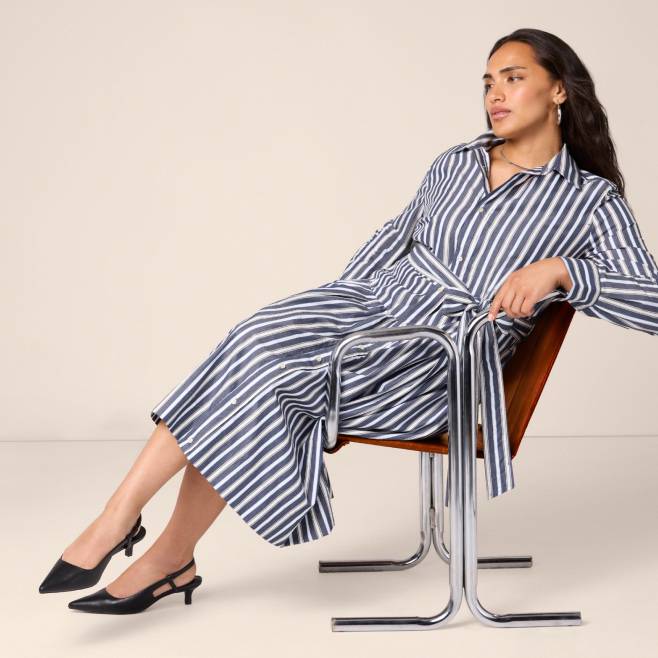 A woman is shown on a chair wearing a belted cotton maxi blue and white striped shirtdress with long sleeves.