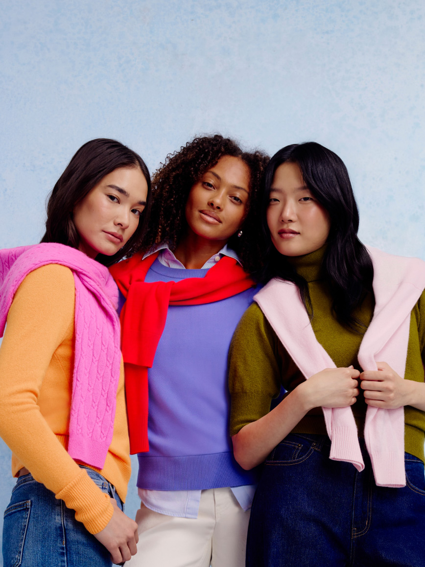 Three adults stand together wearing bright layered sweaters in pink, red, orange, purple and green tones.