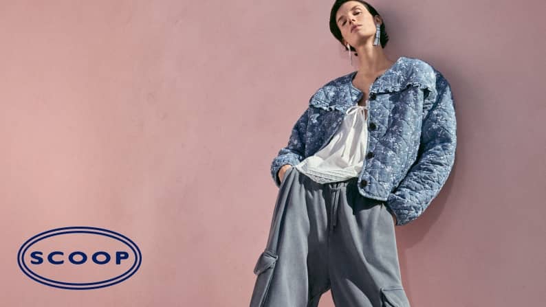 Model wears a blue quilted jacket over a white blouse with gray cargo style pants standing against a soft pink wall.