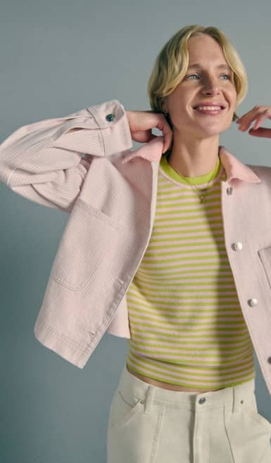 A woman wears a light pink chore jacket over a green striped top, smiling against a grey backdrop.