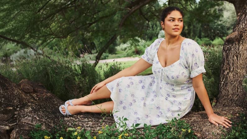 A plus size woman wearing a floral print dress and heels is sitting under a tree.