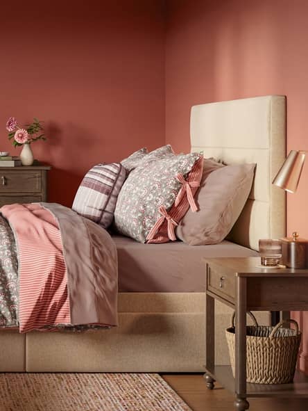 Cozy bedroom with a beige upholstered bed, layered pink and patterned pillows, taupe bedding, and wooden nightstands with decor.