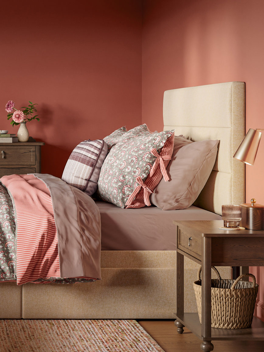 Cozy bedroom with a beige upholstered bed, layered pink and patterned pillows, taupe bedding, and wooden nightstands with decor.