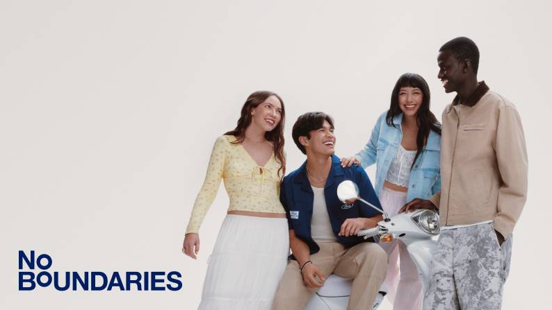 Four young adults smile and laugh around a white scooter, wearing casual spring outfits in a bright studio setting.