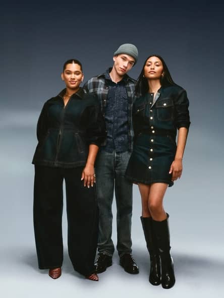 Three adults in dark denim outfits pose together, including a denim dress with tall black boots and wide leg denim pants.