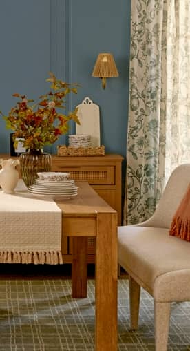A dining room is set with a wooden table and console with decor and dinnerware. A throw lays on a cream upholstered bench.