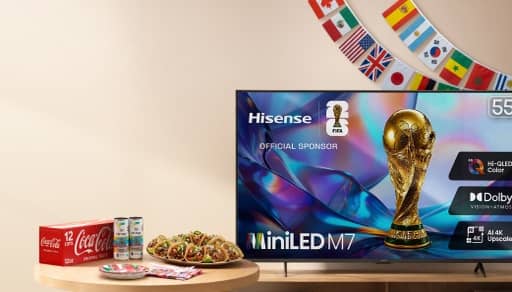 A multi-country flag banner, a TV with a World Cup display, a twelve pack of Coke, two cans of Celsisus, paper plates and napkins and plate of tacos.