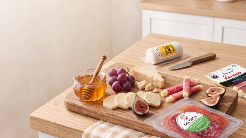 Charcuterie board with cheese, grapes, figs, rolled meat and cheese snacks, honey jar and packaged sliced salami on a wooden table.