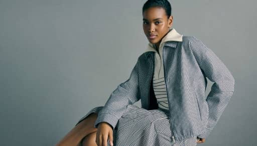 A fashion model wears a checkered jacket and pants over a striped top, seated against a gray background.