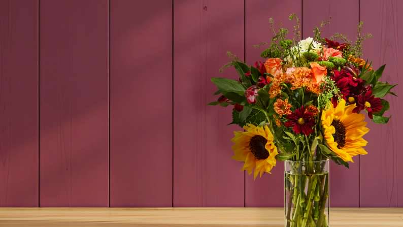 A clear vase of red, orange, and yellow flowers with some green filler on a wood counter against a mauve wood background.