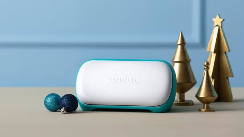 A blue Cricut Joy is playfully framed by mini golden Christmas trees and matching blue ornaments.