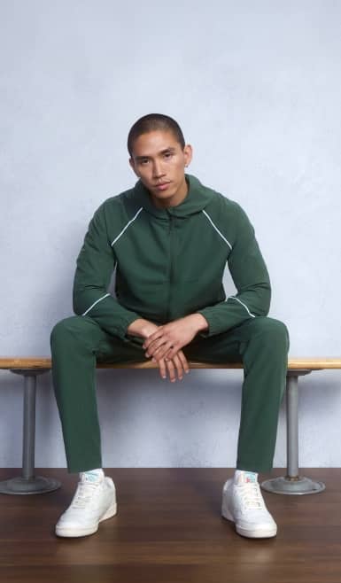 Man sitting on a bench wearing a forest green zip-up active hoodie, green sweatpants and white sneakers in a studio.