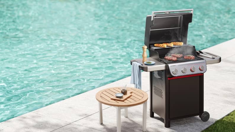 A grill with steaks cooking, with a pool in the background.