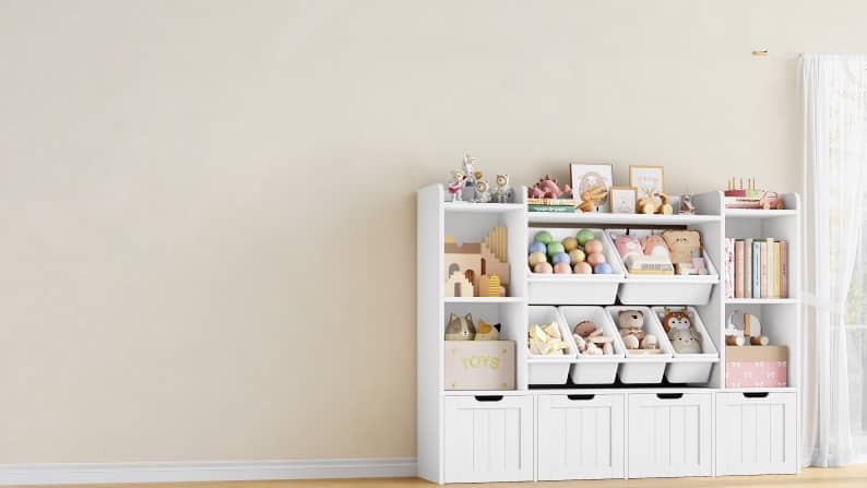 White kids storage shelves with cubbies and bins neatly filled with toys, books and plush animals beside a window with sheer curtains.