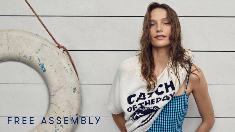 Woman in blue-and-black gingham bathing suit layered with ‘Catch of the Day’ sweater, standing by wall with nautical life preserver.