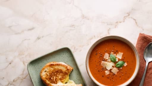 Bowl of tomato soup topped with cheese and basil beside a grilled cheese sandwich on a plate with a spoon nearby.