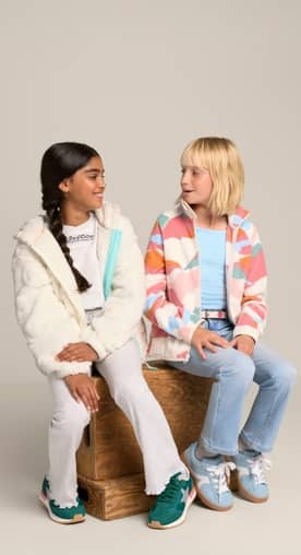 Two little girls wearing colorful fleece are pictured sitting down on a brown block in front of a beige backdrop.