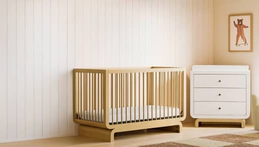 Wooden crib in bedroom with teddy bear in the corner against a dresser.