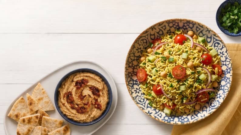 A platter of hummus and pita bread next to a large bowl of long-grain rice with tomatoes, chickpeas, red onions, cucumbers, and herbs.