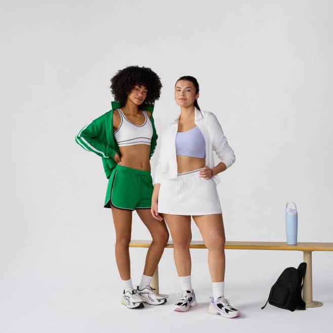 A woman wearing a green track jacket and shorts is standing next to a woman wearing a white track jacket and skirt in front of a bench.