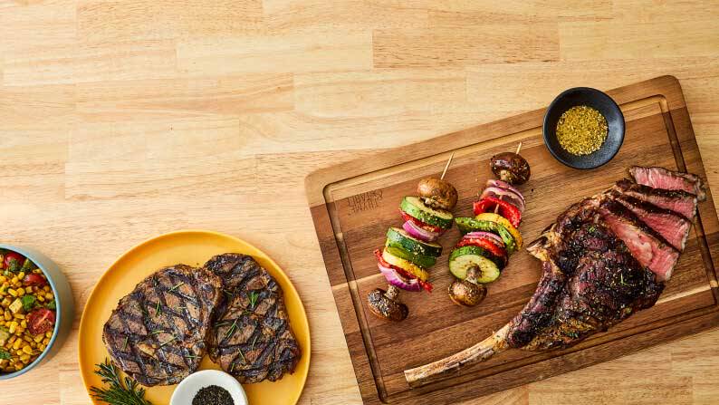 Grilled steaks on plates and a wooden cutting board with sliced tomahawk steak, vegetable skewers, a bowl of seasoning and a side salad.