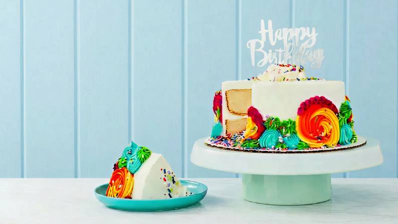 A colorfully iced vanilla cake that has a topper reading “Happy Birthday” sits on a cake server on a table with a plated serving of cake.