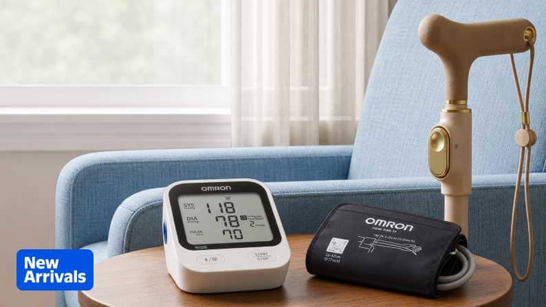 New Arrivals badge. Omron digital blood pressure monitor and cuff on a wooden table beside a beige walking cane in a living room.