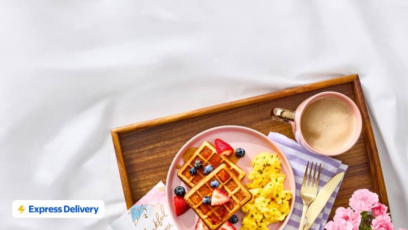 Express Delivery. A wooden tray with a greeting card, flowers, coffee, and a plate of scrambled eggs and waffles topped with syrup and fresh berries.