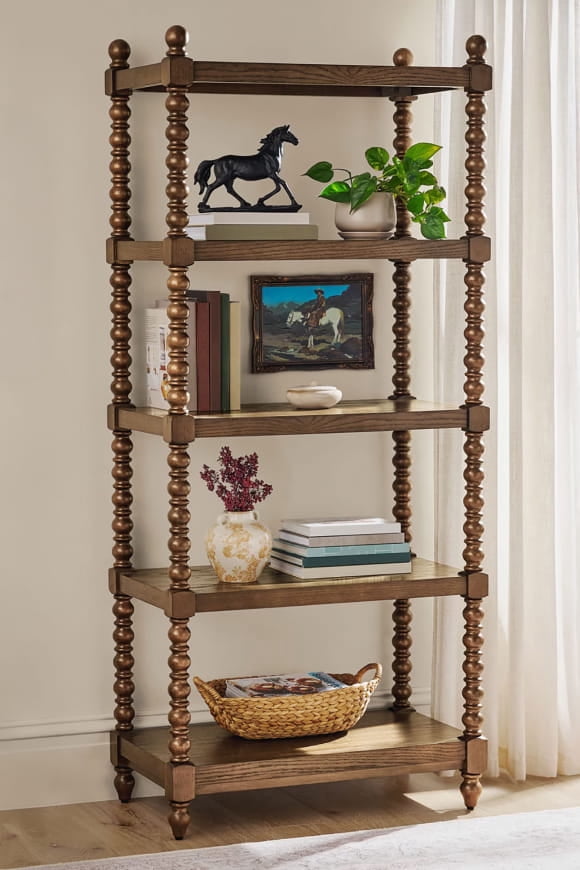 Tall wooden bookcase with spindle legs holding books, framed art, a horse figurine, plants, a vase and a woven basket.