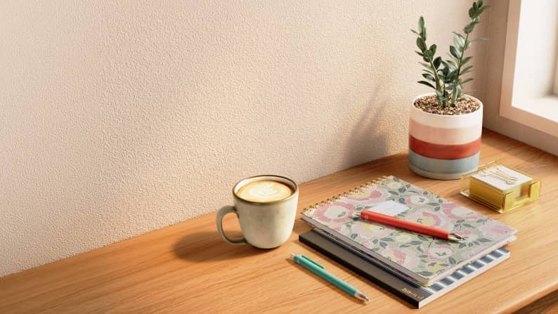 Latte in a ceramic mug beside patterned notebooks, colorful pens, a striped plant pot and a box of paper clips on a wooden desk.