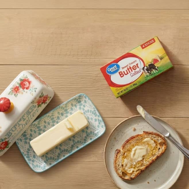 A butter dish with a stick of butter on it next to a box of Great Value salted butter and a plate with a piece of buttered toast on a wooden tabletop.