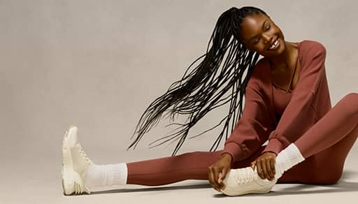A woman wearing a brown set is pictured stretching in cream running sneakers on a beige backdrop.