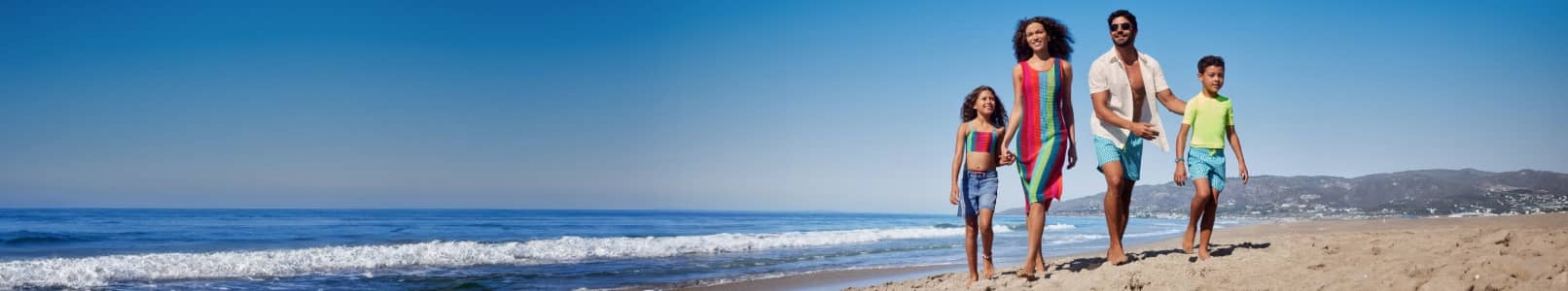 A family of four is shown walking on a beach wearing matching swim styles, from suits to coverups.
