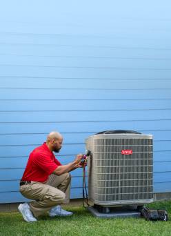 A person in a red shirt and khaki pants, kneeling on green grass while working on a large HVAC system.