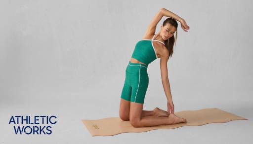 Athletic Works logo in the bottom left corner. A woman is wearing a green active set is posing on a tan yoga mat.