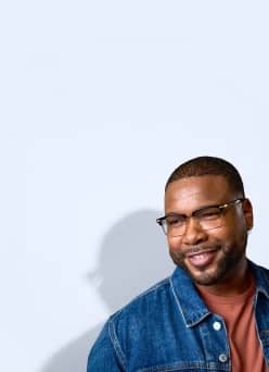 Adult wearing prescription glasses and a denim jacket smiles slightly off camera.