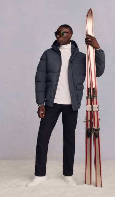 A man is shown wearing a black puffer jacket over a white turtleneck sweater, black jogger sweatpants and polarized flip up shield sport sunglasses.