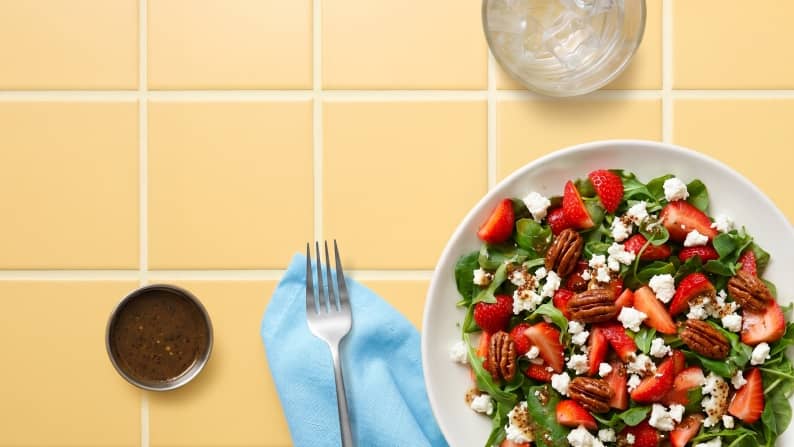 A plate of fresh greens, pecans, strawberries, and crumbled cheese on a tile surface next to a glass of ice water and a small cup of dressing.