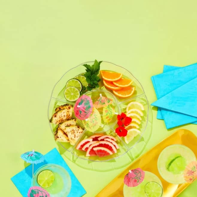 Colorful party supplies featuring a platter, napkins and a plate with slices of fruit.