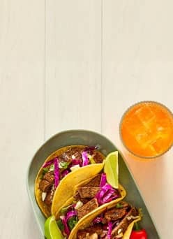 Corn tortilla tacos filled with steak, red cabbage, and onion on a plate garnished with lime wedges next to a glass of orange soda on ice.
