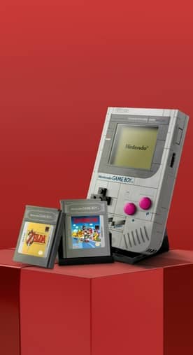 A LEGO-built Nintendo Game Boy is displayed with two game cartridges on a red geometric surface.