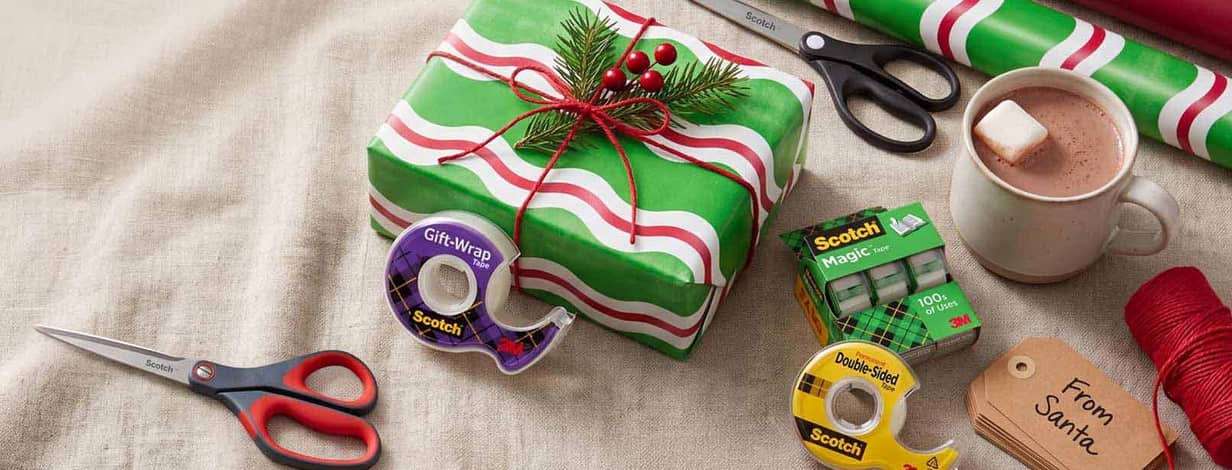 Scotch tape and gift wrap supplies around a wrapped present on a table with hot chocolate and a small card that reads: From Santa.
