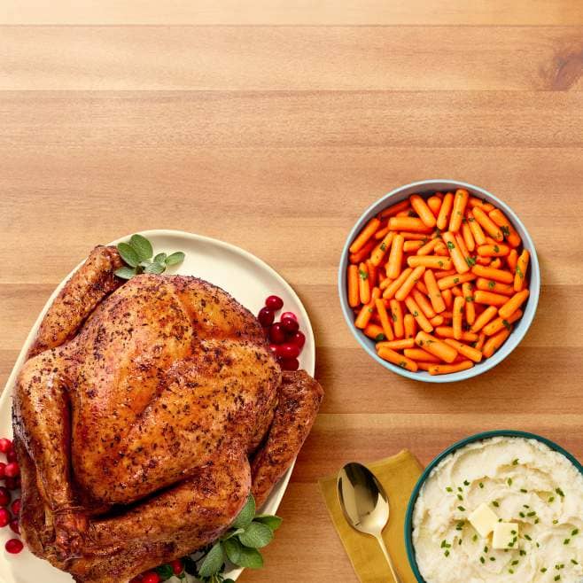 A bowl of mashed potatoes, a roasted turkey on a platter garnished with cranberries and a bowl of baby carrots and herbs are shown.