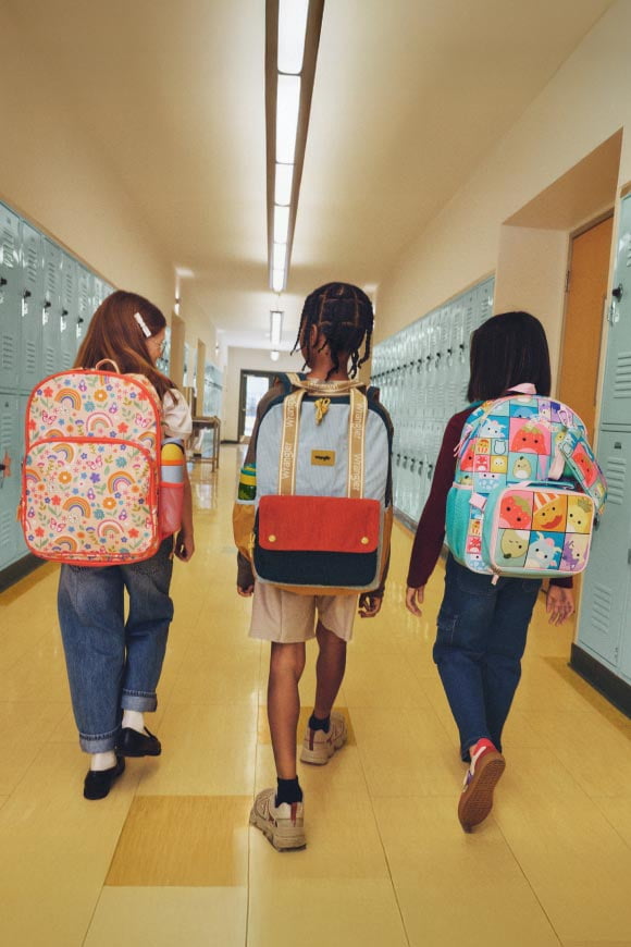 Three kids shot from behind wearing colorful backpacks walking down a school hallway.