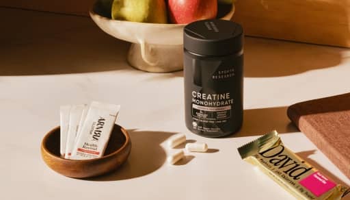 Sports Research creatine monohydrate container with white capsules, Arrae supplement sachets in a dish and a David protein bar on a counter.