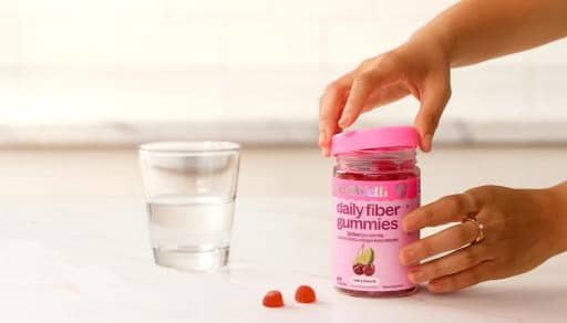 Adult hand opens daily fiber gummies jar beside a glass of water and two red gummies on a countertop.