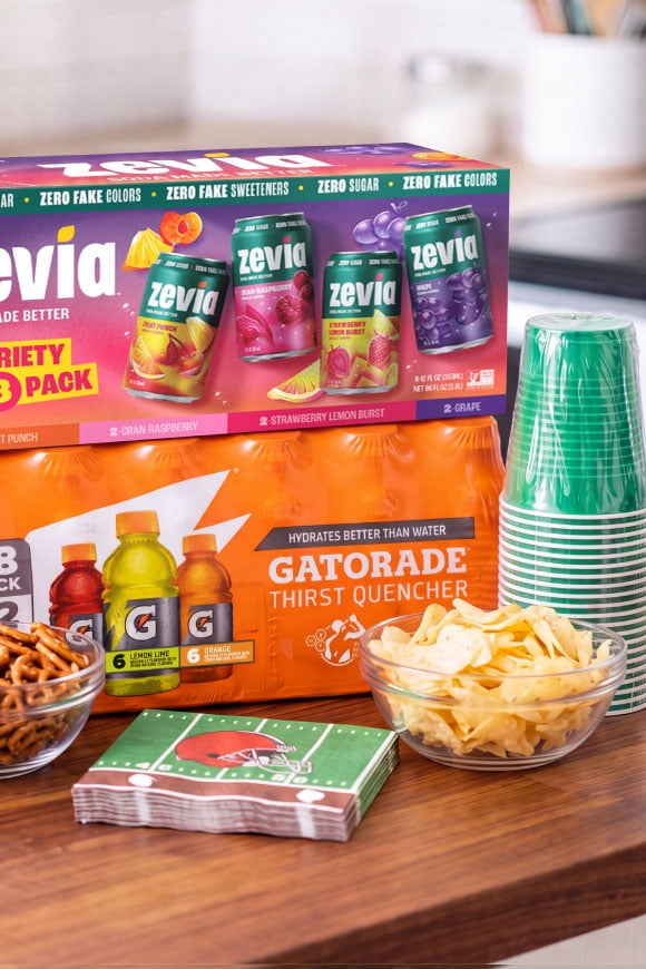Variety packs of Zevia and Gatorade on a wood counter next to chips, pretzels, plastic cups and football-themed napkins.