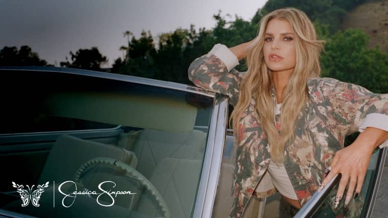 Jessica Simpson logo in the bottom left corner. Jessica Simpson is wearing a printed shirt standing outside of a convertible.