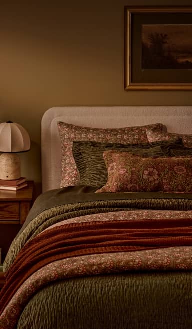 Cozy bedroom with layered green and rust bedding, floral pillows, a bedside table with lamp and books, and framed wall art above the bed.