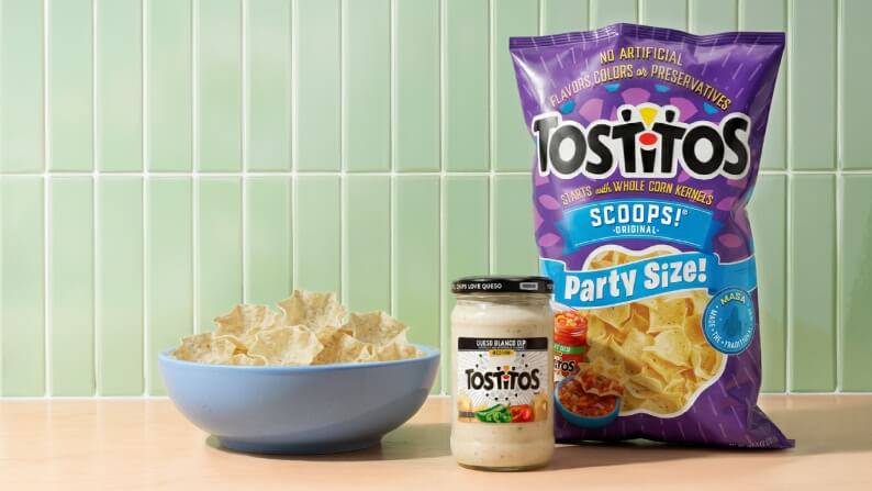 A party size bag of Tostito’s tortilla chips and white queso dip sit on a kitchen counter.
